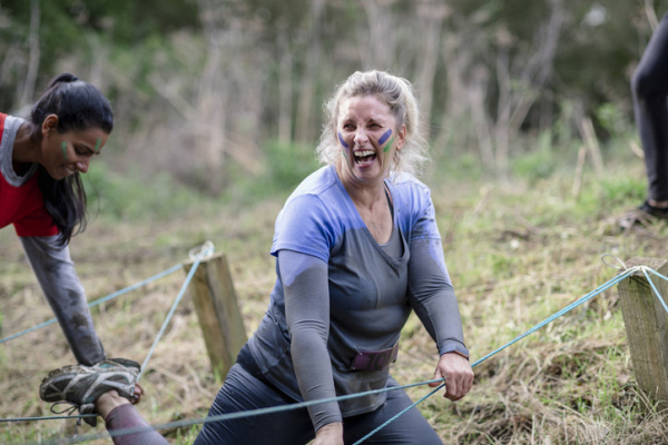 A muddy woman, laughing, goes through mud run obstacle course as a woman leans forward to help; both have colorful stripes on cheeks